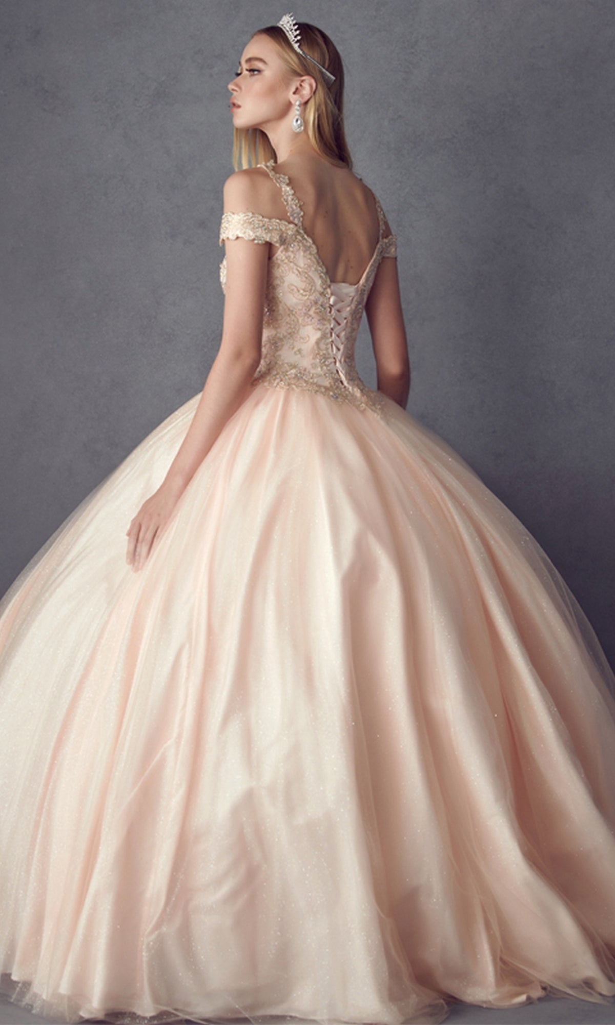 Wearing the Juliet 1430 Cold-Shoulder Glitter Quinceanera Dress, a woman stands against a gray background, her light pink gown featuring a fitted lace bodice and full tulle skirt as she faces slightly away.